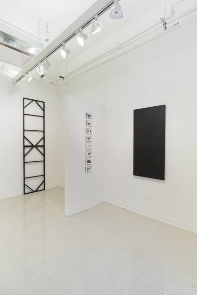 Overview of the installation of artworks by Cameron Richie. This includes a metallic structure on the left side, a series of geometric paintings stacked on the well in the center of the image, and a black painting with geometric shapes.