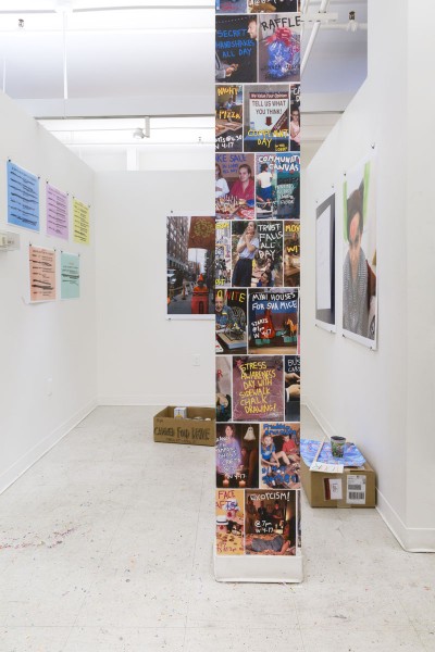 Installation view of 30 Days: An Attempt At A Community with a column covered in print in front and a different print on a white wall in the background by Cassidy Toner.