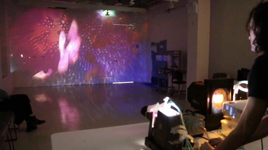 A person handles a light source and an image projector, casting a large scale image projection on the wall in front of him of a specular light with small dots on blue and violet background