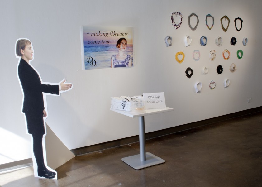 Installation view of a person cut-out from cardboard, a table with white shirts, a print on the wall with a person and an ocean backdrop, and different colored shirt collars