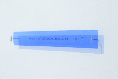 A translucent blue sign mounted to a white wall with metal T pins which reads, “What is an embodied existence for you?”