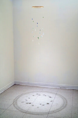 A mobile, hung from the ceiling by a thin clear thread, featuring various tiny glass sculptures of microbes casting a dynamic shadow on the floor.