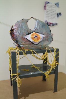 A wire sculpture with a large central eye placed on top of a pedestal, draped in a sheet of wet SCOBY.