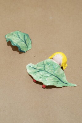 A small clay sculpture of a figure lying underneath a leaf.