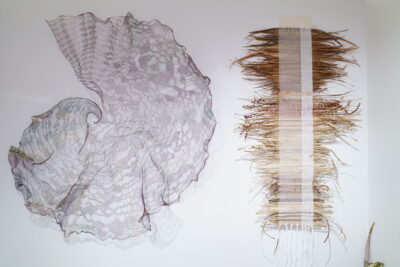 An intricate, spiraling wire sculpture hung against a white wall beside a weaving made with natural materials.