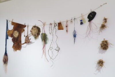 A group of weaving, wire sculptures and art objects made of various natural materials hung on a line against a white wall.