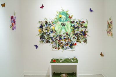 A collage featuring a painted portrait of a woman surrounded by images of butterflies and plant life hanging on a white wall, between two smaller collages on opposite walls and above a small installation resembling a mossy altar.