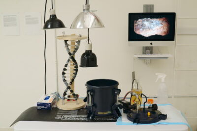 Miscellaneous laboratory materials and equipment arranged across a tabletop, in front of a monitor showing an image of a mineral.
