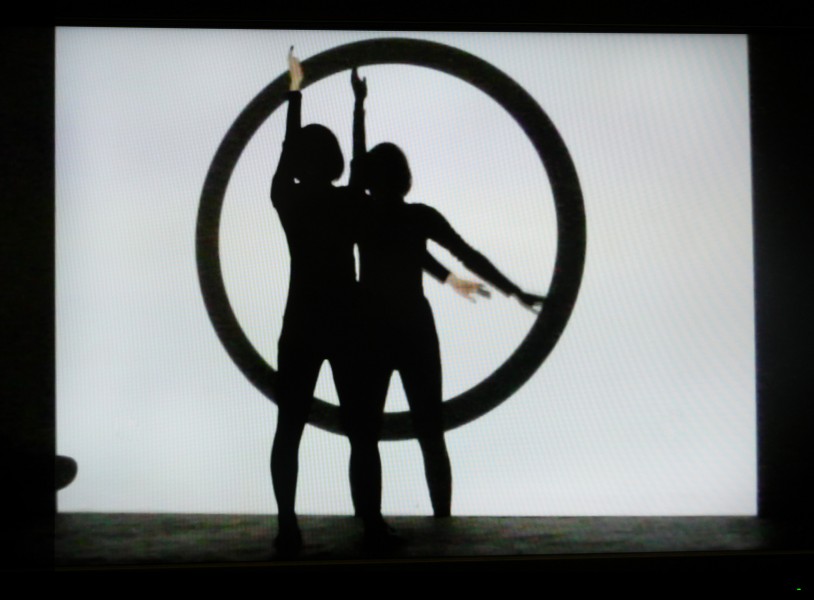 A person standing in front of a wall with a projection of a black circle on a white background with a hand raised and the other hand indicating 4 o'clock position on the circle.