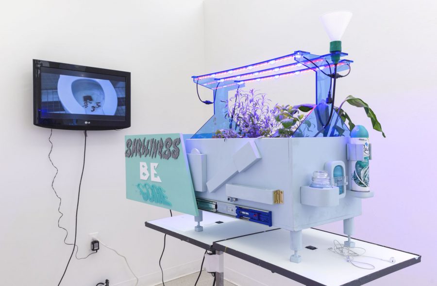 Installation shot of a sculpture with grow lights that has feet, and says Smelliness be Gone, with a video on the wall.