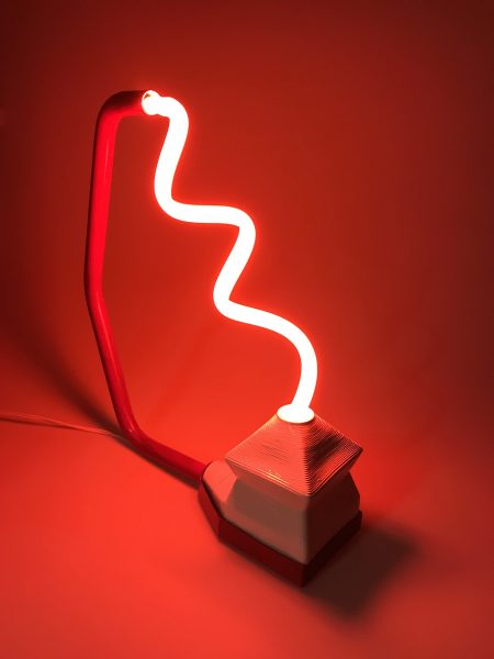 A sculpture by Dakin Platt. The sculpture is has a rod and base holding a thin twisted neon light. The color of the light is bright red. The background is a plain backdrop.