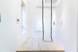 Installation view of multiple paintings and a sculpture by Dana Avendano. Abstract images with different tones of white and black and geometric shapes. Large wooden platform in the center of the space and two long ropes of hair hanging from the ceiling.