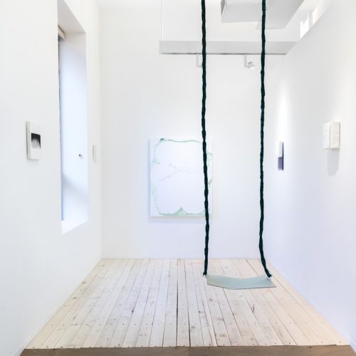 Installation view of multiple paintings and a sculpture by Dana Avendano. Abstract images with different tones of white and black and geometric shapes. Large wooden platform in the center of the space and two long ropes of hair hanging from the ceiling.