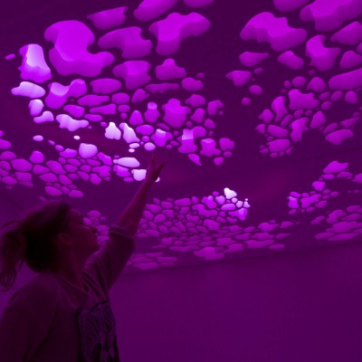 A person with their left hand raised to the ceiling. The ceiling is illuminated in purple, and it is formed with organic shaped sculptured in it.