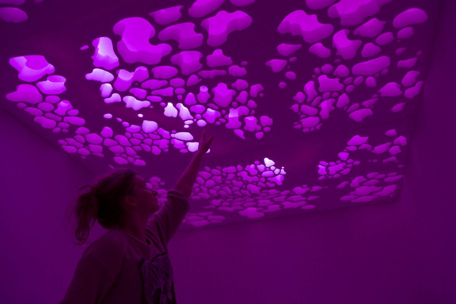 A person with their left hand raised to the ceiling. The ceiling is illuminated in purple, and it is formed with organic shaped sculptured in it.