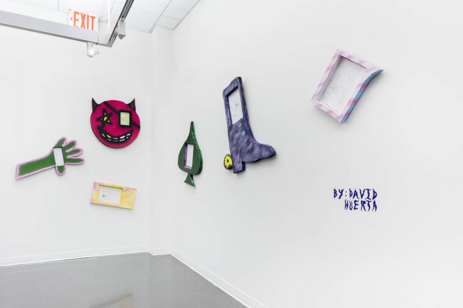 Installation view of sculptures made of organic materials, painted in green, red, yellow, blue, and pink, with various shapes, from a hand, a boot, a red evil emoji shape, to a rectangle frame with baby pink and blue. On the far right side is a text written By David Huerta.