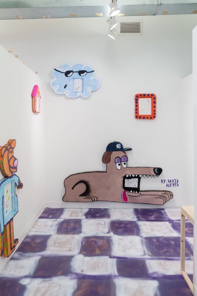 Installation view of a dog sculpture made of plain board material colored in brown, sitting with an object in his mouth, a blue cloud installed on the same wall above the doc, an orange frame on the right on the same wall, a figure with a pig head and blue shirt and orange legs ion the left wall and the floor is painted with blue and white squares