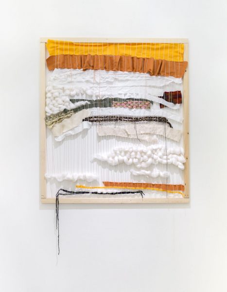 Wall mounted loom with yellow, brown, and white wool and fabric woven throughout.