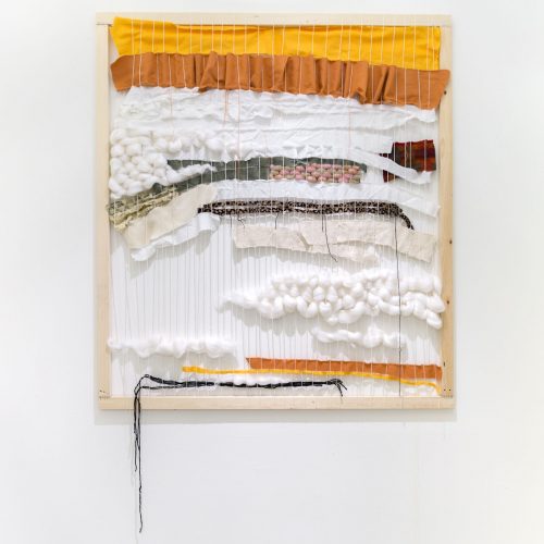 Wall mounted loom with yellow, brown, and white wool and fabric woven throughout.