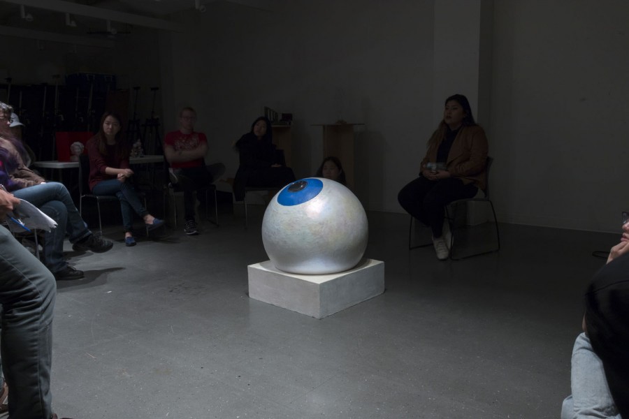 Installation view of an organic blue eye sculpture in a room full of people sitting on chairs in the dark with the eye being  the only thing illuminated in the room