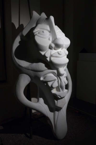 Sculpture made from white organic material with rounded and curved shapes, illustrating a face with distorted features like big eyes, a nose bulbed at its base and going over the head in the middle of the forehead, prominent cheeks, etc., being illuminated from the upper right side casting shadows on the left part of the frame