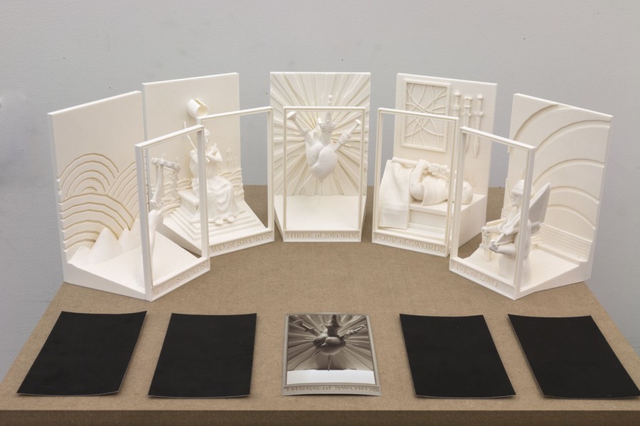 Five white sculptures placed on a plywood board with five cards in front of them, representing abstract, rounded, organic shapes in a rectangular frame