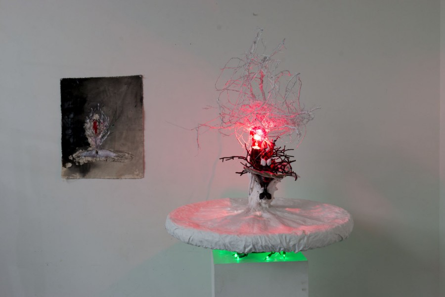 Installation of an abstract sculpture with white branches rising, and black branches at the bottom with a red light in the middle, and on its left side is an abstract painting of a horizontal object with something coming out of it looking like the sculpture