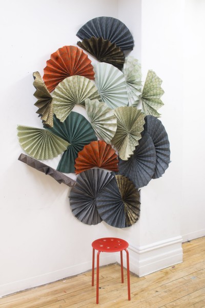 Installation of hand fans made of papers in blue, olive, and orange colors, mounted on a corner with a red chair under the installment