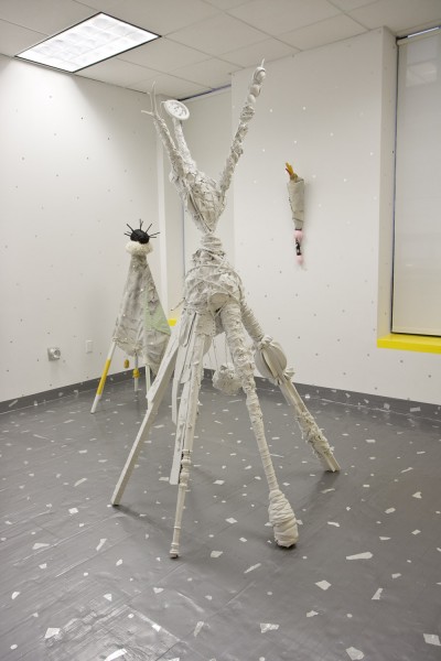 Installation view of three sculptured with organic shapes, two installed on the floor with a plastic foil and white angular shapes on it, and one sculpture is installed on a white wall with silver dots on the wall