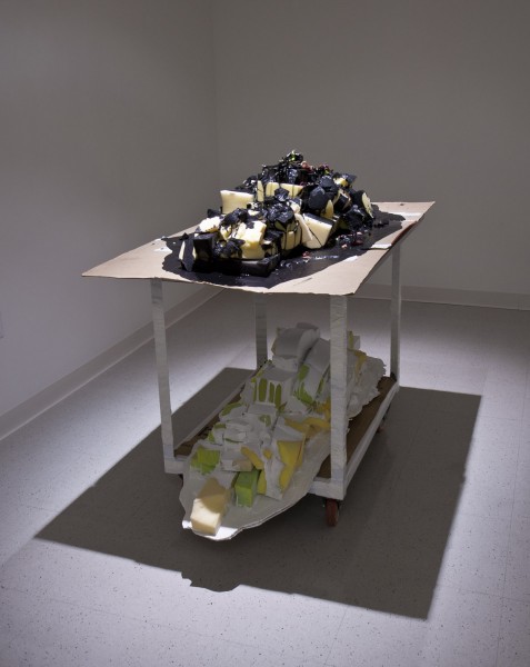 Installation of sculpture made of sponges installed on a tabletop and the table bottom shelf. The pile of the sponge on the tabletop has black painting dripping on over, and the pile of sponges on the bottom shelf has white paint dripping on it.