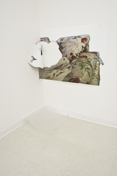 A print mounted on the wall near the corner of a sofa with a sofa pillow, a blanket, and a cut-out in the shape of a person from under the blanket, and a small mirror on the bottom left corner.