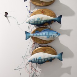 Four fishes sculptures with blue and white colors are installed on the wall, each with a wooden piece and linked together with wires in different colors. Two of the fishes are mounted on the wall upside-down.