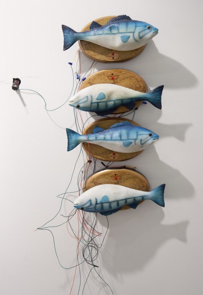 Four fishes sculptures with blue and white colors are installed on the wall, each with a wooden piece and linked together with wires in different colors. Two of the fishes are mounted on the wall upside-down. 