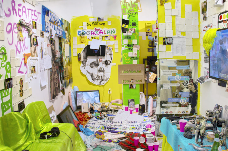Multi-media installation with sections of walls painted yellow and green with paper stuck to the way and text in paint marker. A television monitor is visible on the left wall and a video is being shown. A couch covered in neon green velvet is across from the television. The space is has papers additional television monitors and pink plastic cups.