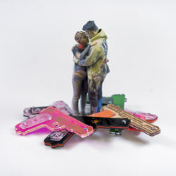 3D print in plaster of two people, dressed in modern casual clothing, embracing. They stand in a pile of laser-cut acrylic and wood handgun shapes in various bright colors.