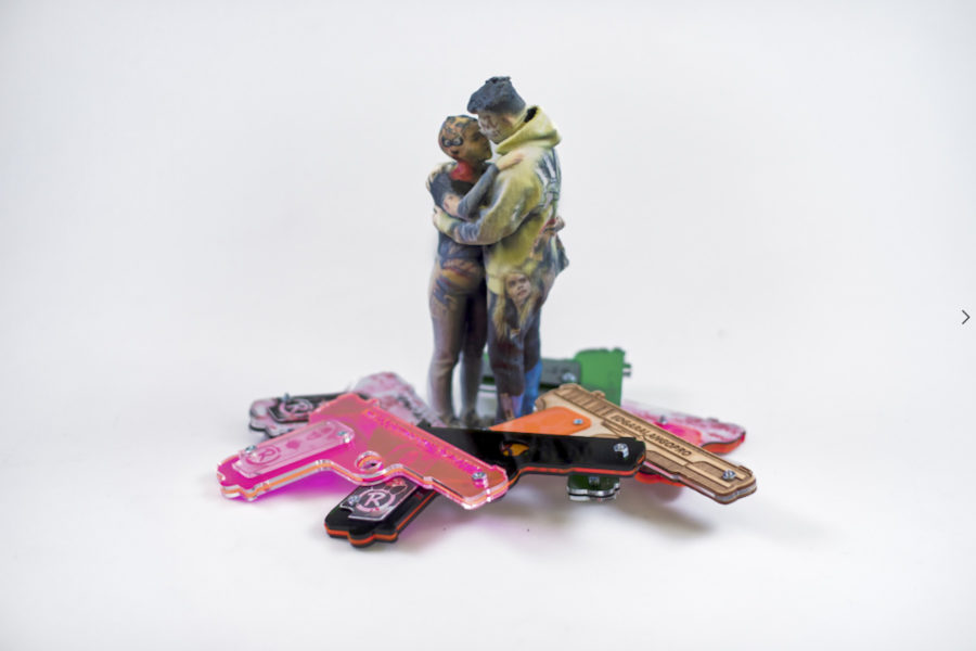 3D print in plaster of two people, dressed in modern casual clothing, embracing. They stand in a pile of laser-cut acrylic and wood handgun shapes in various bright colors.