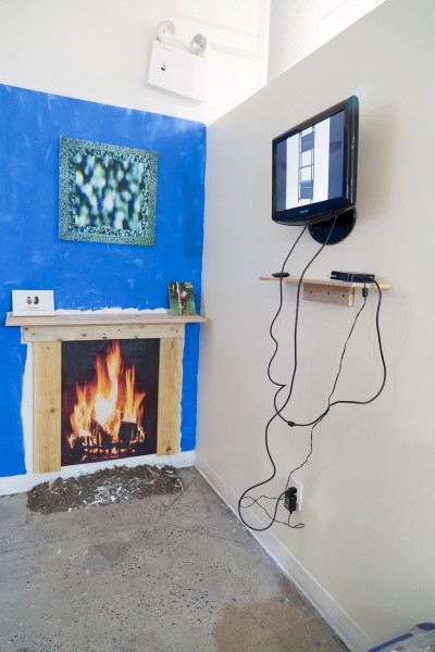 A corner of a room with a fake fireplace with a print of a fire, a wooden frame, two photos, a fake mirror on a blue wall, and a TV beside with an image on it.