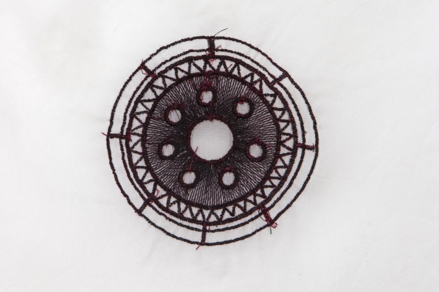 Embroidered  red circle-shaped on a white piece of fabric, with small vertical lines, angled lines, and circles going to the center of the larger circle