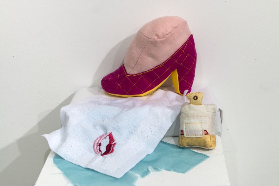 Installation of feet in a red high heel shoe made of fabric placed on a white piece of cloth with a red lips shape on a side and a perfume bottle made of material in white and yellow.