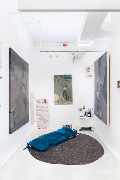 Installation of painting by Emily LaRosa. Expressionistic paintings of figures in various colors. Text written on large sheets of paper, a table with various items on it, a blue sleeping bag. 