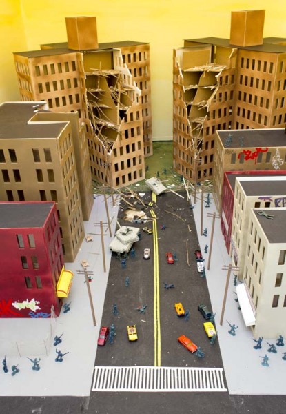 Bird-eye view of a street under siege with soldiers on the streets, cars, red, white, and beige buildings, and two taller buildings each with a corner destroyed