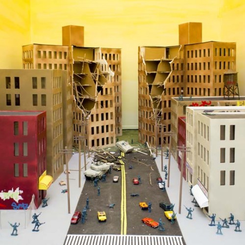 Miniature model of a city street under siege with soldiers in the streets, cars, a tank, red, white, and beige buildings, and two tall buildings each with a corner destroyed