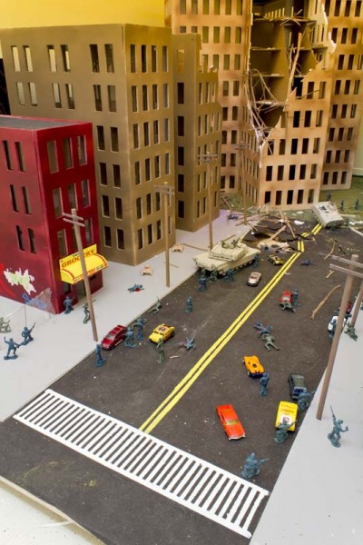 Miniature model of a city street with cars, people, soldiers, and a tank, an intact red building, two other buildings, and one building with a corner on the top destroyed