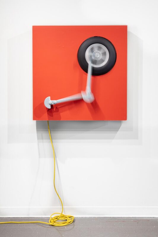 An artwork by Titus McBeath titled "Cain and Able (Cain)" consisting of a red painting with a kinetic sculpture mounted on it with a spinning wheel. A yellow extension cord hangs down from the painting and is coiled on the floor. 