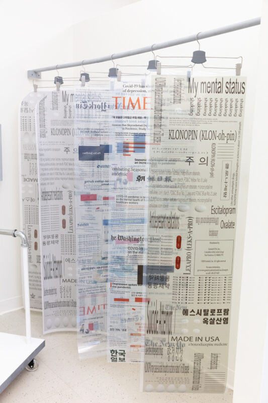 Typography in color and black and white digitally printed on translucent white sheets are suspended from a white pole using metal pants hangers.