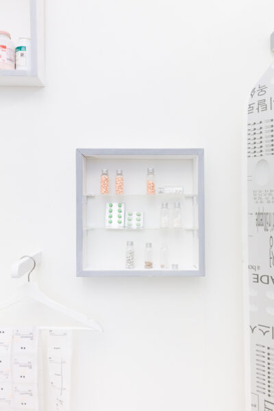 Various stylized packages of pills sit on glass shelves inside a minimalist cabinet mounted on a white wall.