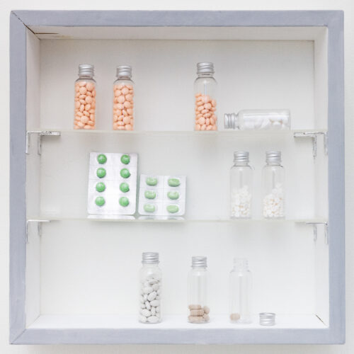 Pills of various sizes and colors in unlabeled glass vials and blister packs are arranged on glass shelves in a small wooden cabinet painted white with a grey frame.