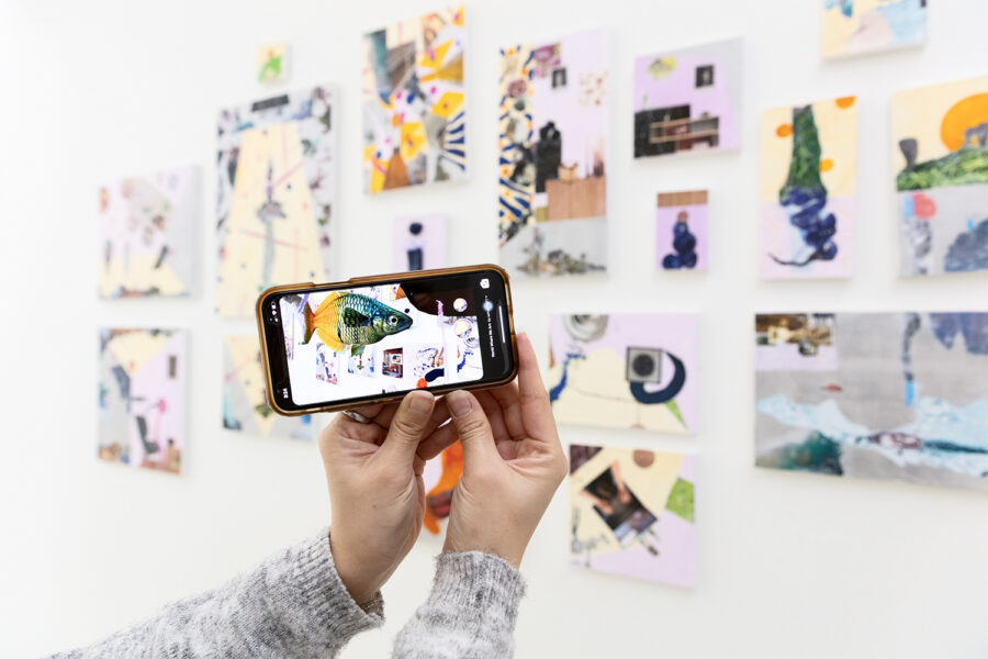 Two hand hold an iPhone in front of an installation of small acrylic paintings mounted on a white wall. On the iPhone screen we see a fish hovering in front of the paintings, demonstrating the Augmented Reality component of the installation.