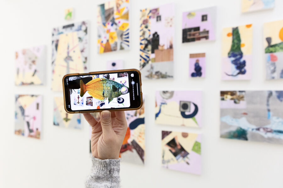 A hand holds an iPhone in front of an installation of small acrylic paintings mounted on a white wall. On the iPhone screen we see a fish hovering in front of the paintings, demonstrating the Augmented Reality component of the installation.