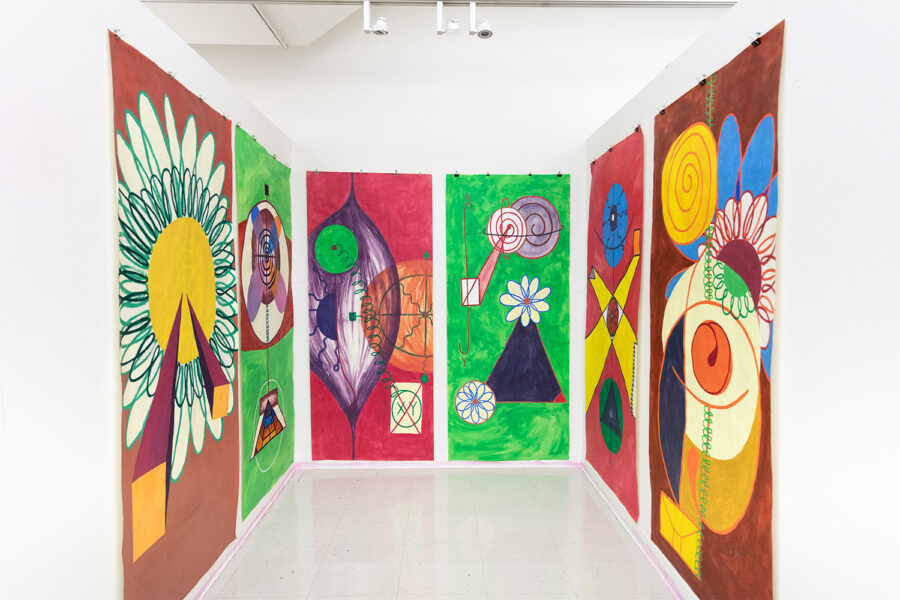 An installation view of six large, colorful paintings with geometric abstractions featuring images of eyes and flowers hanging in a white-walled studio.